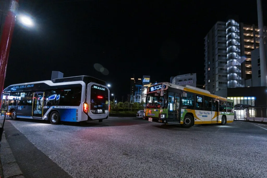 Two buses at a night-time Ichinoe bus stop with illuminated interiors and surrounding street lights.