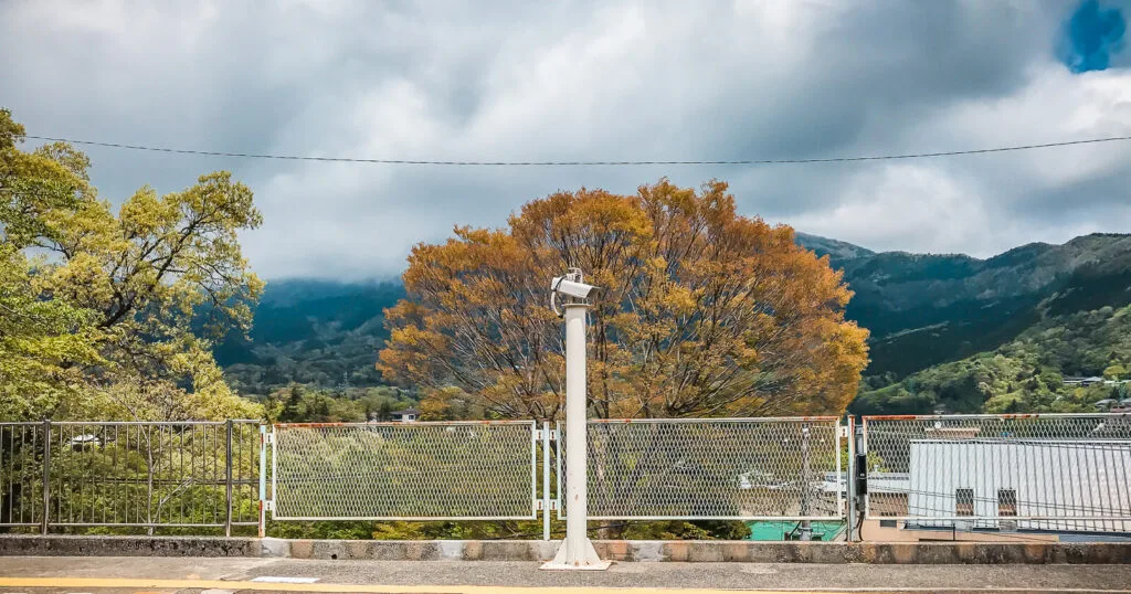 View from a station platform in Hakone, showcasing a vibrant autumnal tree against a backdrop of misty mountains and a partly cloudy sky.