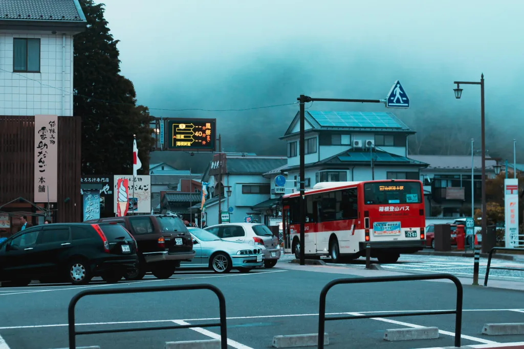 A red bus with destination signage in Japanese characters at a bus stop near Lake Ashinoko, Hakone, with misty mountains in the background and various vehicles parked alongside.