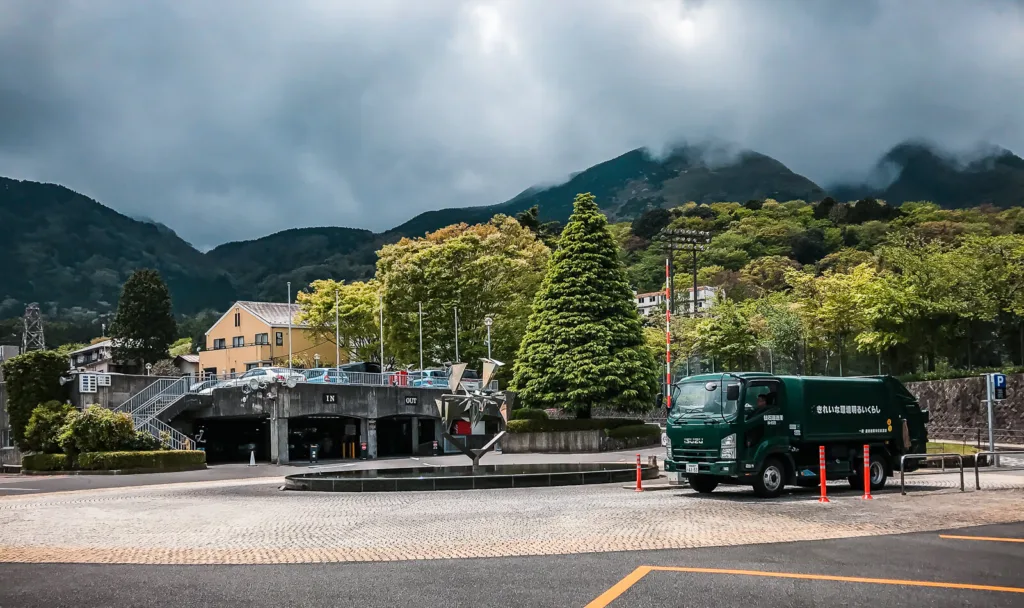 View of the Open Air Museum at Hakone with a green truck parked in the foreground, a paved plaza, and misty mountains in the background.