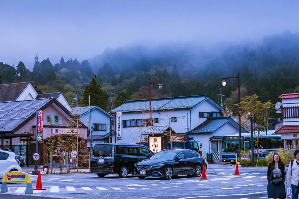 A scenic view of a street near Lake Ashinoko in Hakone with misty mountains in the background, traditional Japanese architecture, and pedestrians.
