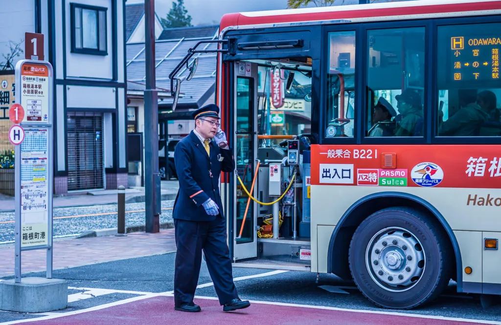 A bus conductor in uniform saluting beside a red and white bus at a station near Lake Ashinoko, Hakone.