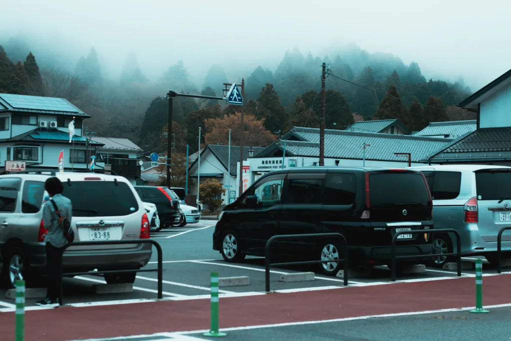 Misty mountain backdrop behind a street scene in Hakone, with vehicles parked and a pedestrian crossing the road.