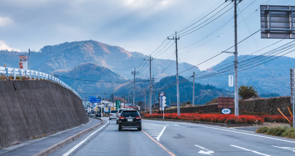 A straight road with a few vehicles, leading towards distant mountains under a slightly cloudy blue sky in autumn. The view is captured from inside a car traveling from Tokyo to Nikko.