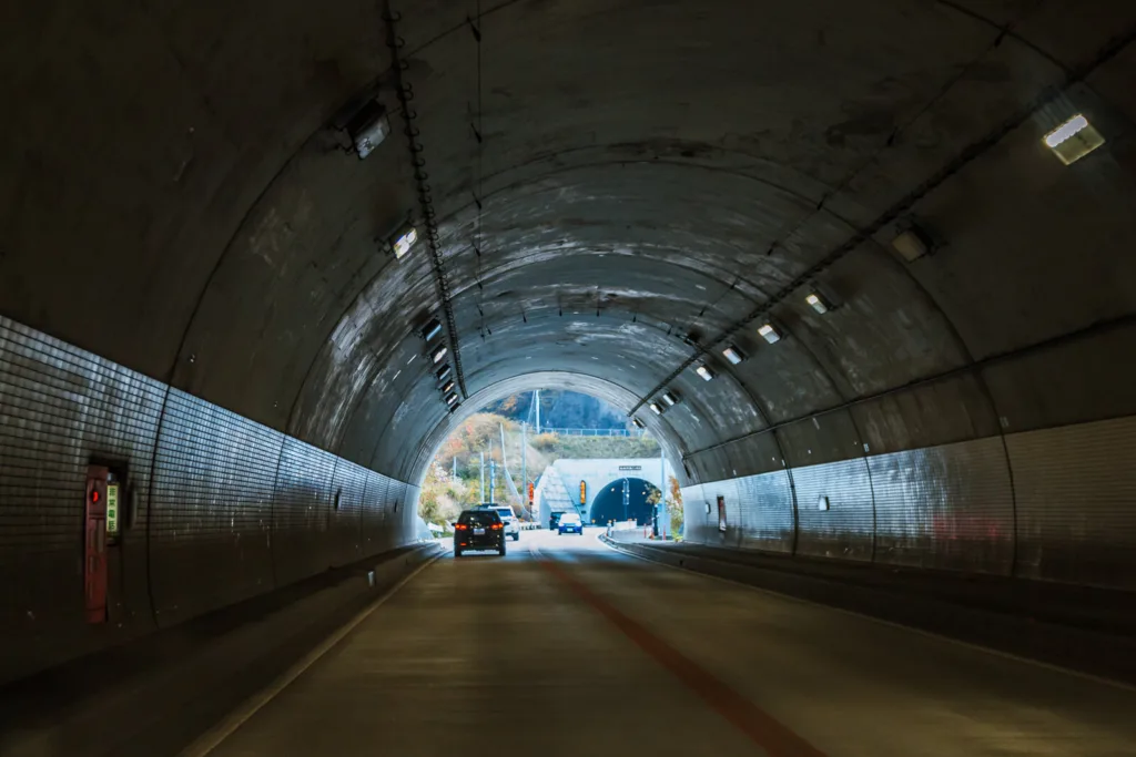 A straight road passing through a tunnel with a few vehicles, showing bright light at the other end. The scene is set during autumn and captured from inside a car traveling from Tokyo to Nikko.