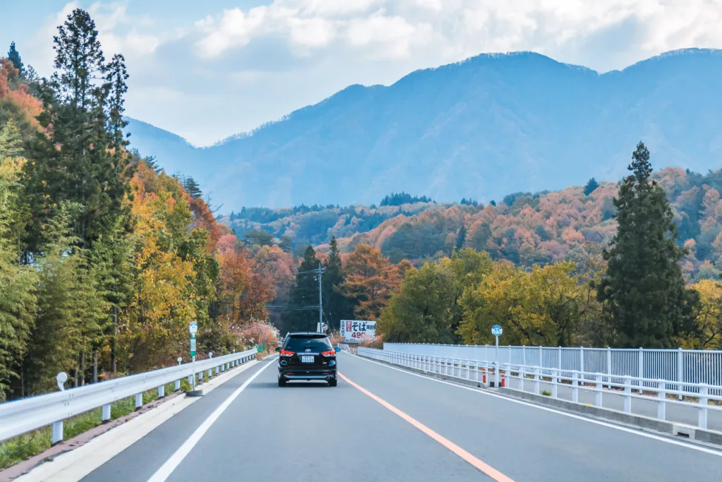 A straight road with a few vehicles, leading toward mountains under a slightly cloudy blue sky in autumn. The photo is taken from inside a car traveling from Tokyo to Nikko.