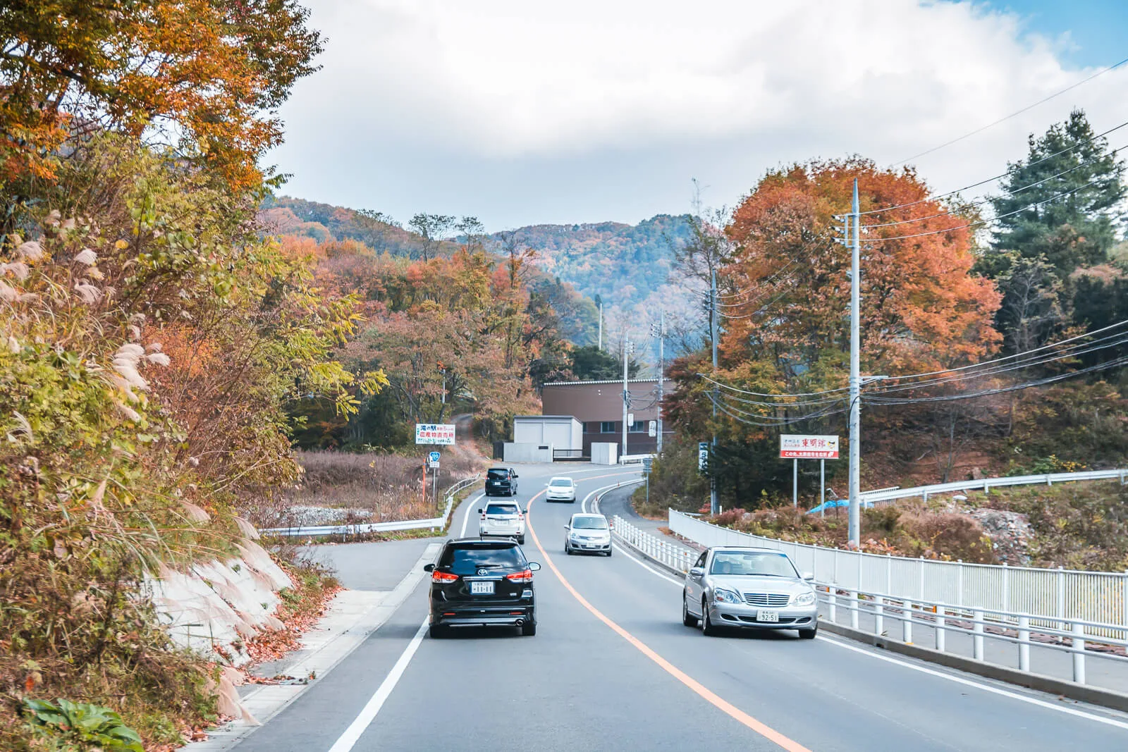 A straight road with a few vehicles, leading toward distant mountains under a slightly cloudy blue sky in autumn. Captured from inside a car during a journey from Tokyo to Nikko.