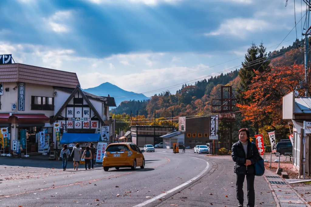 Autumn leaves scattered around the roads of Nikko with a few people and cars visible. In the background, mountains rise under a cloudy sky with rays of sunlight breaking through.