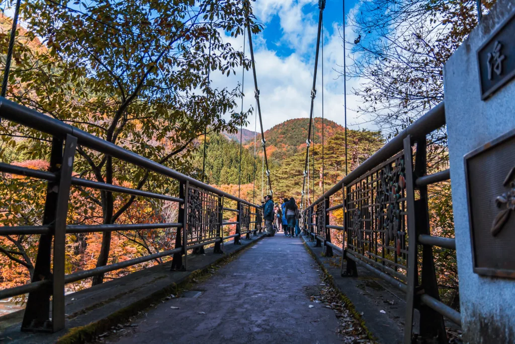 A low-angle photo taken from one end of a bridge near Fukiware Falls in Nikko, showing the entire length of the bridge with tourists on it and vibrant autumn leaves in the background.