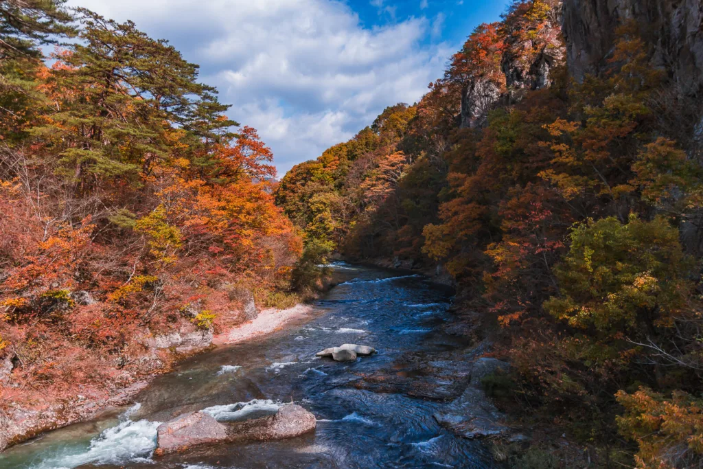 A view from the bridge near Fukiware Falls in Nikko, showing the river flowing below, surrounded by vibrant autumn leaves, with mountains and a partly cloudy sky in the background.