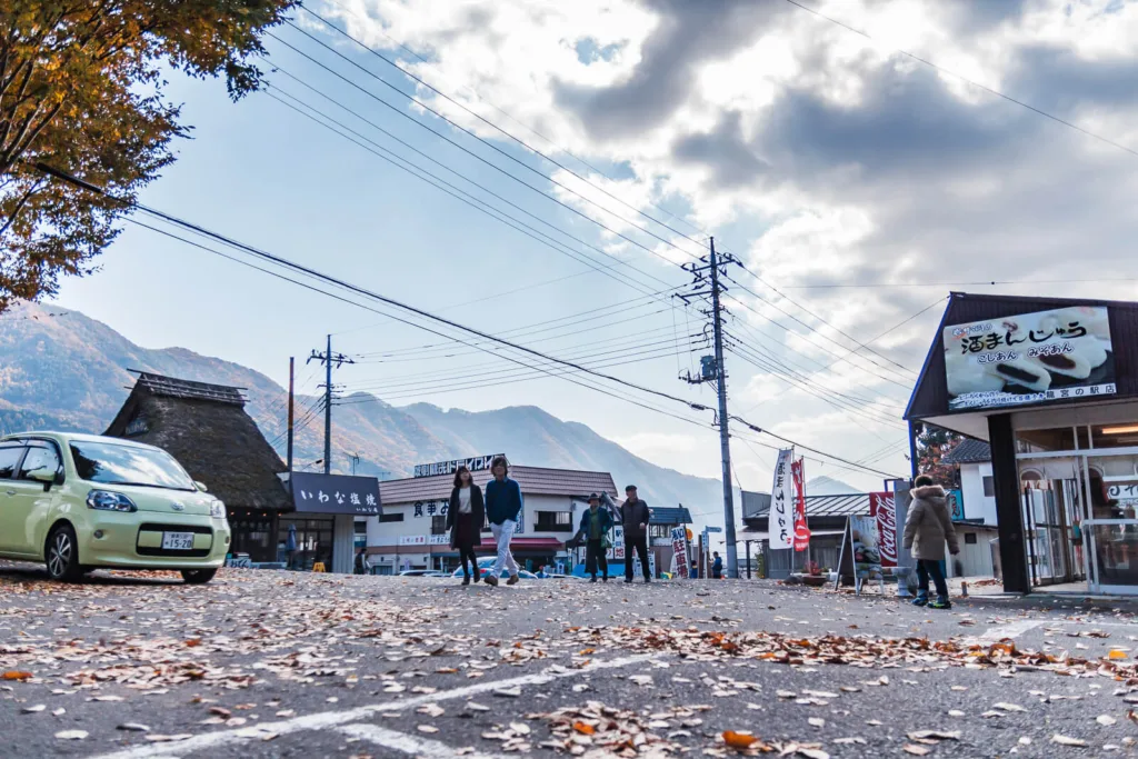 A parking area near the entrance to Fukiware Falls in Nikko, with autumn leaves scattered around, a road, shops, mountains, and a blue sky with some clouds in the background.