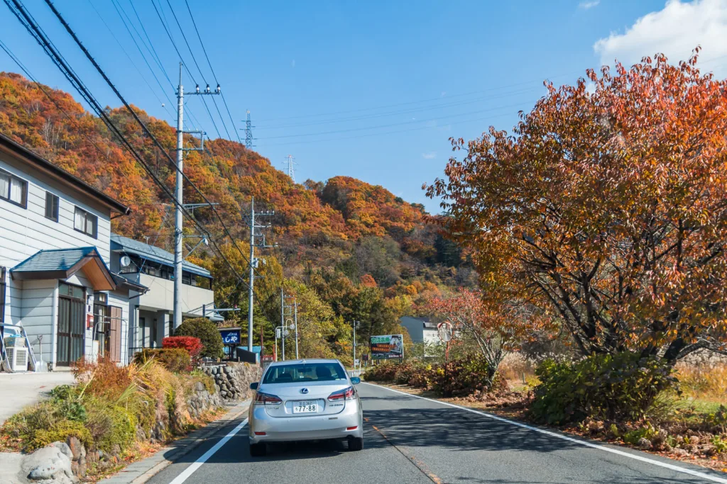 A road leading towards mountains with a few vehicles, houses on the side, and a slightly cloudy blue sky in the background. The photo is taken from inside a car traveling from Tokyo to Nikko during autumn.