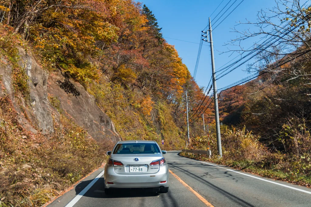 A road stretching ahead with a few vehicles, mountains in the distance, and a slightly cloudy blue sky, taken from inside a car traveling from Tokyo to Nikko during autumn.