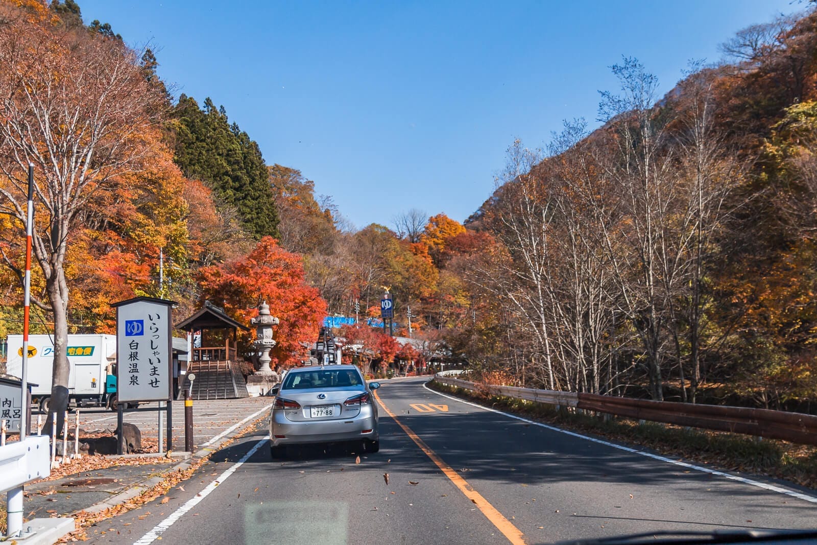 A road stretching ahead with a few vehicles, cemetery on one side, mountains in the distance, and a slightly cloudy blue sky, taken from inside a car traveling from Tokyo to Nikko during autumn.