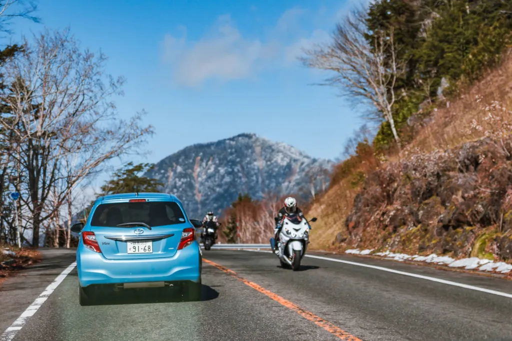 A road with a few vehicles, mountains ahead under a slightly cloudy blue sky, and patches of snow on the roadside. The photo is taken from inside a car traveling in Nikko during autumn.