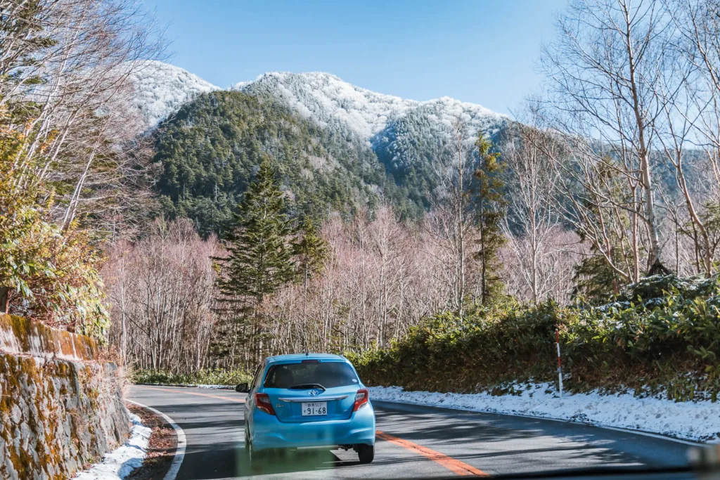 A winding road with a few vehicles, slightly snow-capped mountains in the distance, and patches of snow on the roadside under a blue sky. The photo is taken from inside a car traveling in Nikko during autumn.