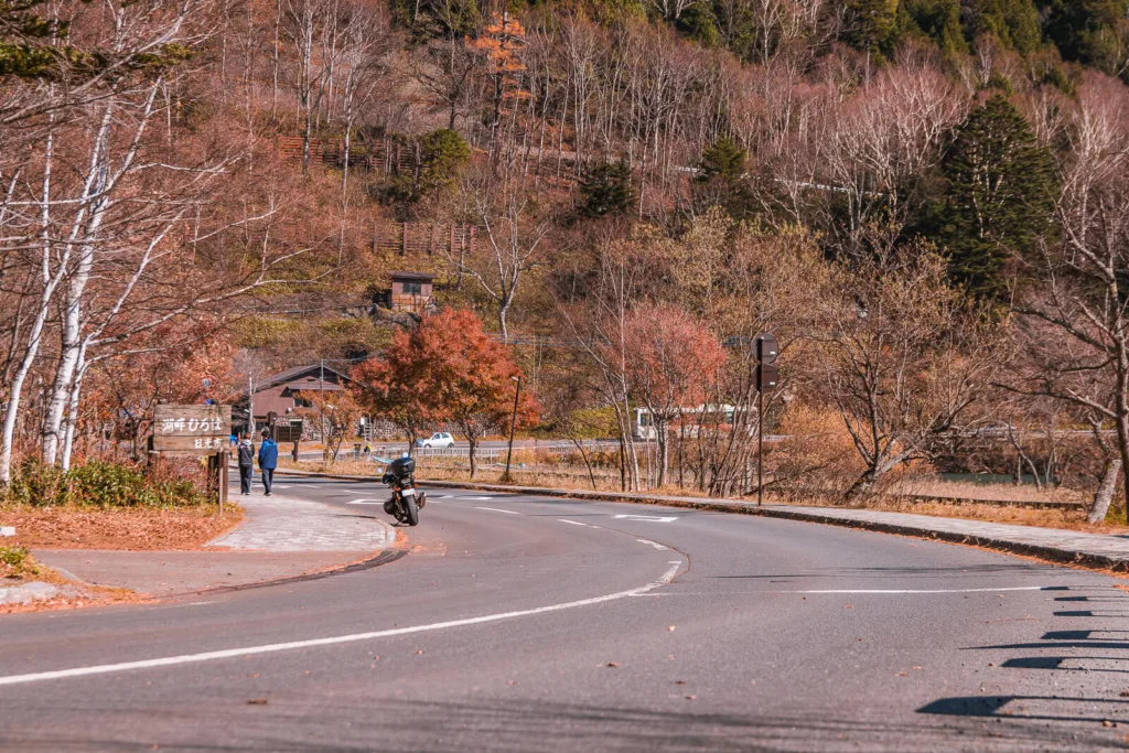 A winding road near Lake Yunoko during autumn, with a few people, a bike, and vehicles including a car and a bus in the distance under a clear daytime sky.