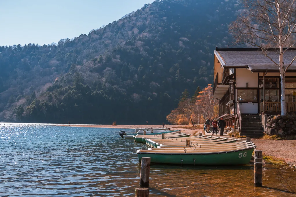 A view of Lake Yunoko during autumn, with the Kyukamura Nikko Yumoto hotel on the right side and rowing boats parked along the lake's edge during daytime.