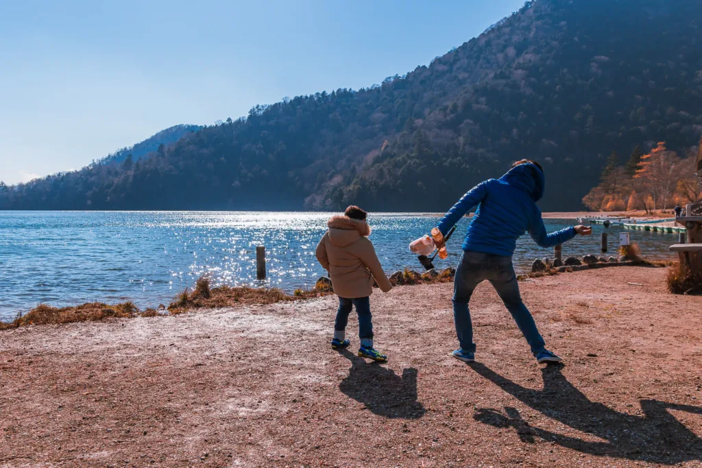 A kid and a man enjoying their time at Lake Yunoko during autumn, with parked rowing boats and a mountain in the background on a sunny day.