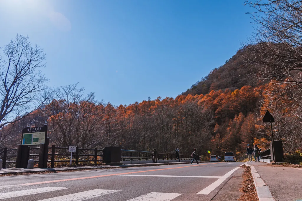 A road near Lake Yunoko with a few people and vehicles, surrounded by autumn colors and a mountain in the background on a sunny day.