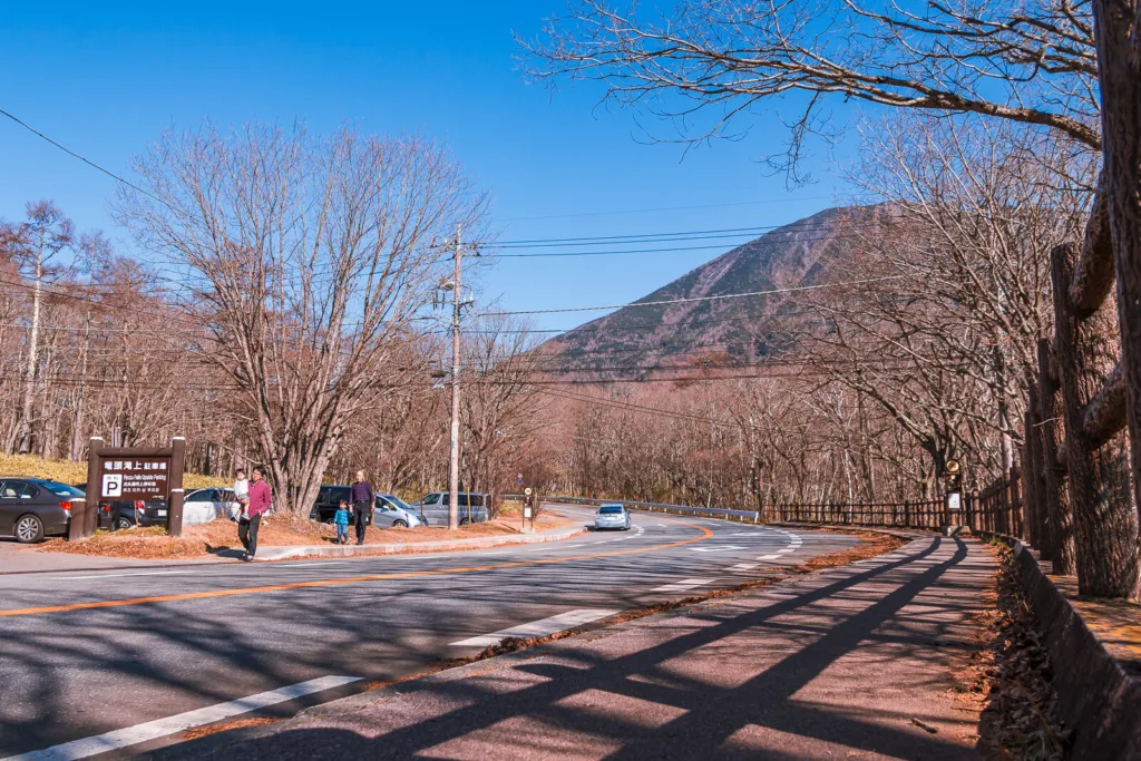 A winding road near Lake Yunoko with a few people and vehicles, surrounded by vibrant autumn colors and a mountain in the background under a sunny sky.