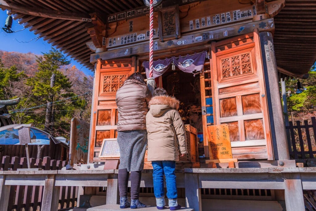 A mother and son praying to the Dragon-Headed Kannon statue near the entrance area of Ryuzu Falls, surrounded by autumn foliage.