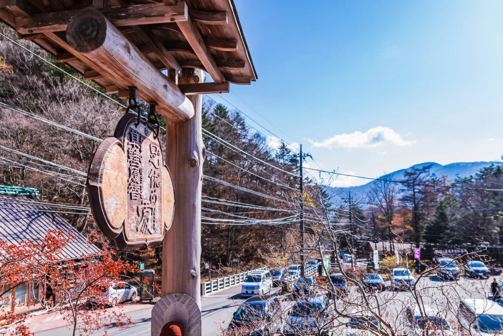 A foot rest area in front of Ryuzu Kannon Temple in Nikko, featuring a board with Japanese writings, a parking area, and mountains under a clear sky in the background.