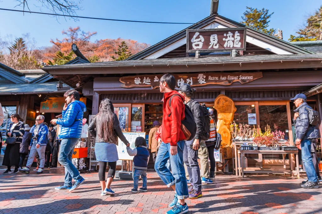 The entrance to the viewing platform of Ryuzu Falls, with a souvenir shop and tea house nearby, and people gathered in front of the tea house on a sunny autumn day.