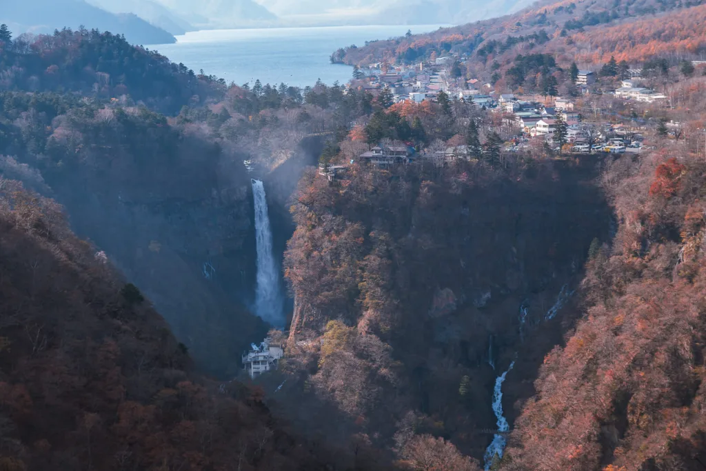 A distant view of Kegon Falls in Nikko, surrounded by autumn foliage under a clear sky.