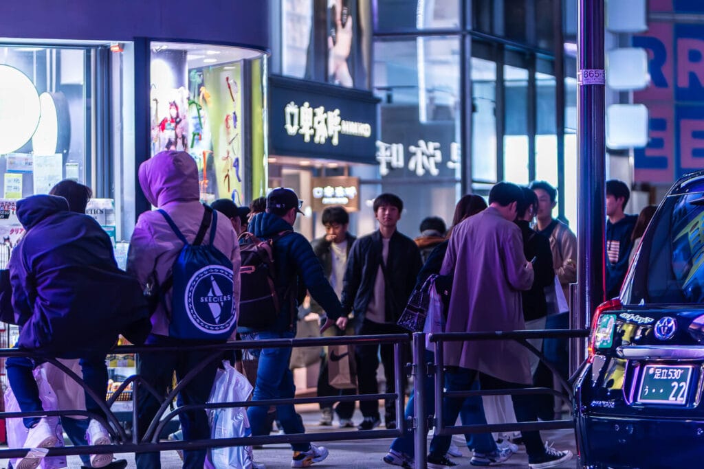 People sitting on railings on the sidewalks of a street in Harajuku at night.