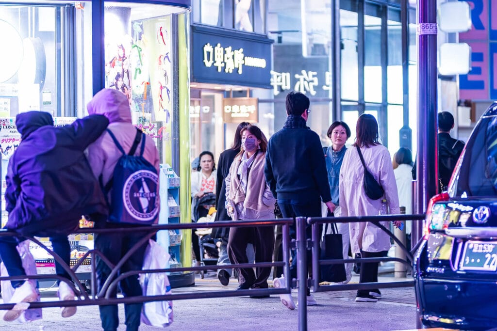 People sitting on railings on the sidewalks of a street in Harajuku at night.